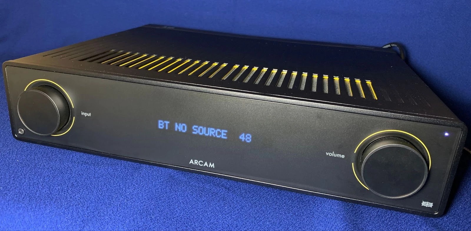 The appearance of the Arcam A15 immediately attracts attention. The Radia series is distinguished by minimalism and restraint: a frosted glass front panel, two large metal encoders, a solid metal case with ventilation holes and yellow accents create a modern and recognizable image.